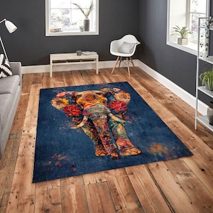 May include: A rectangular blue area rug featuring a colorful elephant design. The elephant is adorned with floral patterns in red, yellow, and orange. The rug is placed on a wooden floor in a room with a window and a white chair.