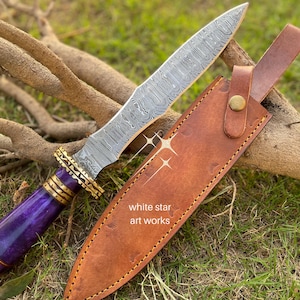 May include: A Damascus steel knife with a purple handle and gold accents, resting on a brown leather sheath. The blade has a unique pattern. The sheath is secured with a brass button. The text "white star art works" is visible.