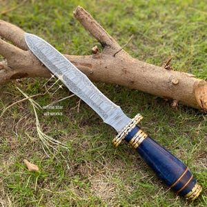 May include: A decorative dagger with a patterned silver blade and a blue and gold handle. The dagger rests on green grass next to a brown log. The text "white star art works" is visible.