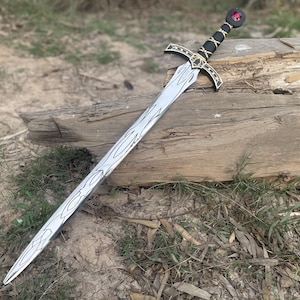 Może przedstawiać: Srebrno-czarny miecz dekoracyjny z ozdobną rękojeścią i ostrzem. Miecz ma czarną rękojeść ze złotymi akcentami i czerwonym klejnotem na głowicy. Ostrze ma wzór słojów drewna. Miecz spoczywa na kłodzie.