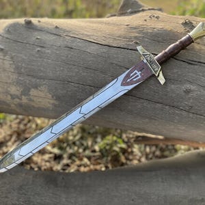Peut inclure: Une épée décorative avec une lame argentée et une poignée enveloppée de cuir marron. La garde est dorée, et la lame présente un motif décoratif et un symbole de trident. L'épée repose sur une bûche en bois vieilli.