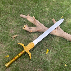 May include: A decorative sword with a silver blade and a gold-colored hilt. The hilt features a cross-shaped design with red and green accents. The sword is lying on green grass next to a piece of wood.