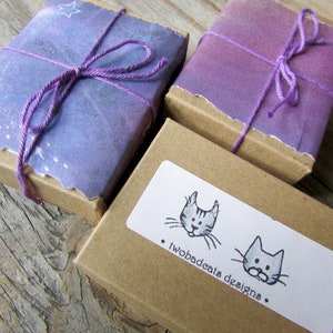 May include: Two small, brown gift boxes with purple and white paper wrapping. The boxes are tied with purple string and have a small, white sticker with a black and white illustration of two cats and the text "twobadcats designs".