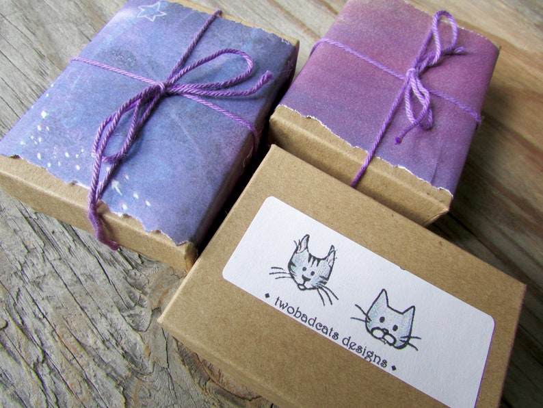 May include: Two small, brown gift boxes with purple and white paper wrapping. The boxes are tied with purple string and have a small, white sticker with a black and white illustration of two cats and the text "twobadcats designs".