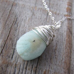 Pu&ograve; includere: Un ciondolo di pietra preziosa blu chiaro avvolto in filo d'argento, appeso a una collana a catena d'argento.