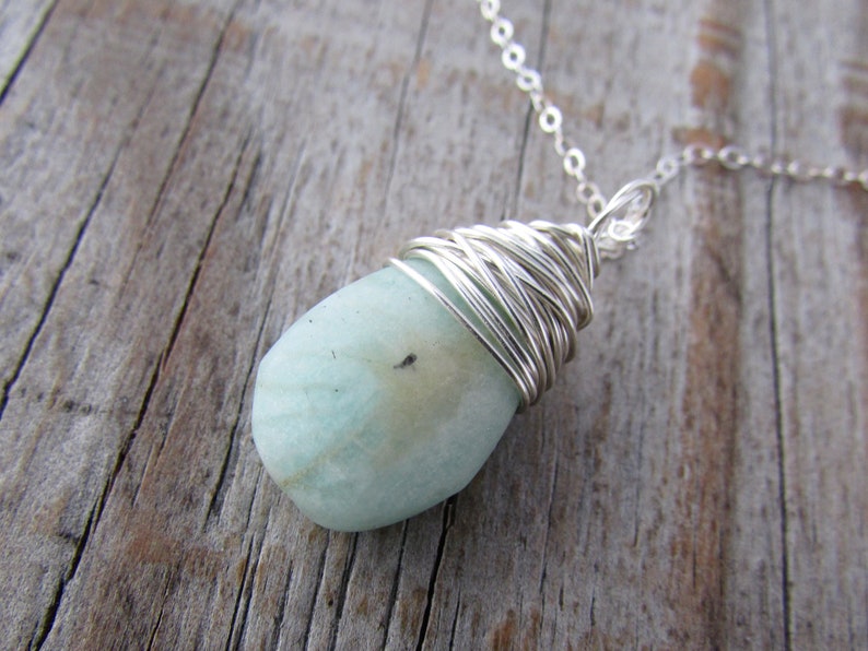 Pu&ograve; includere: Un ciondolo di pietra preziosa blu chiaro avvolto in filo d'argento, appeso a una collana a catena d'argento.