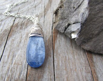 May include: A blue kyanite pendant necklace with a silver chain. The kyanite stone is wrapped in silver wire. The necklace is displayed on a wooden surface next to a gray rock with a crystal.