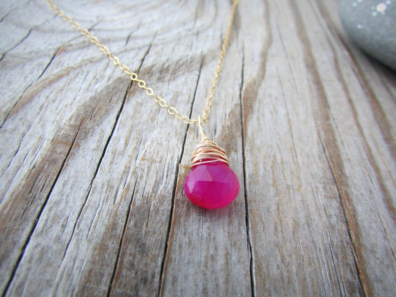 May include: A delicate gold chain necklace with a pink gemstone pendant. The gemstone is wrapped in gold wire and has a faceted design.