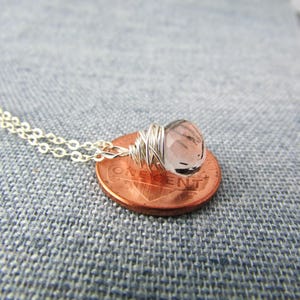 May include: A delicate silver chain necklace with a light pink gemstone wrapped in silver wire. The necklace is displayed on a penny.