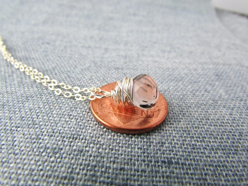 May include: A delicate silver chain necklace with a light pink gemstone wrapped in silver wire. The necklace is displayed on a penny.