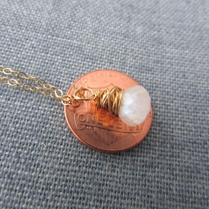 May include: A gold chain necklace with a small white gemstone pendant wrapped in gold wire. The pendant is shown next to a US penny for size comparison.