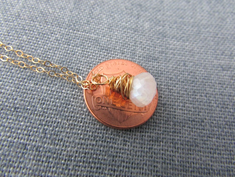 May include: A gold chain necklace with a small white gemstone pendant wrapped in gold wire. The pendant is shown next to a US penny for size comparison.