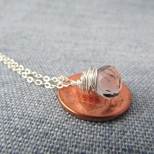 May include: A silver chain necklace with a small, light pink gemstone wrapped in silver wire. The necklace is displayed on a penny.