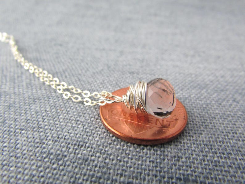 May include: A silver chain necklace with a small, light pink gemstone wrapped in silver wire. The necklace is displayed on a penny.