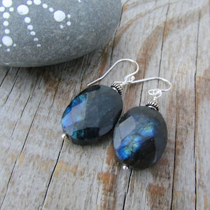 Può includere: Orecchini pendenti in labradorite con ganci in argento sterling. Le pietre ovali sfaccettate mostrano riflessi blu e verdi iridescenti. Gli orecchini sono appoggiati su una superficie di legno invecchiato, con una pietra dipinta sullo sfondo.
