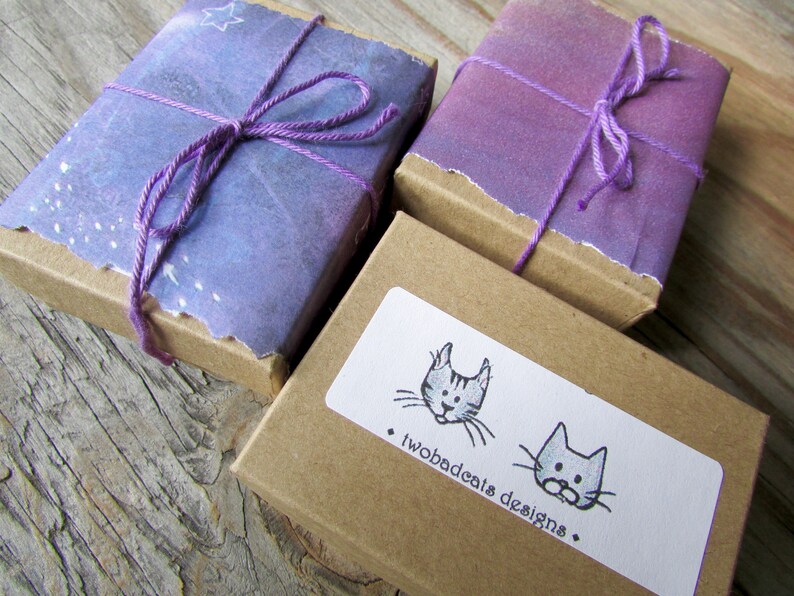 May include: Three small, rectangular boxes wrapped in purple and brown paper. The boxes are tied with purple string and have a white label with a black and white illustration of two cats and the text "twobadcats designs."