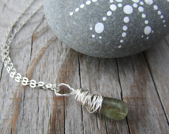 May include: A silver necklace featuring a green gemstone pendant wrapped in silver wire. The pendant hangs from a delicate chain. The necklace rests on a wooden surface, with a gray stone in the background.