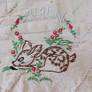 May include: A white fabric with a brown embroidered deer lying down surrounded by green leaves and red flowers. The word "AMEN" is embroidered in blue above the deer.