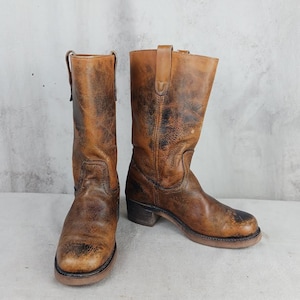 May include: A pair of brown leather cowboy boots with a distressed, worn appearance. The boots have a mid-calf height, a rounded toe, and a small heel. The leather shows dark patches and wear marks, giving them a vintage look.