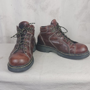 May include: A pair of brown leather ankle boots with black soles and yellow-laced black laces. The boots have a classic design with visible stitching and metal eyelets. The boots are a rugged style, suitable for outdoor activities.