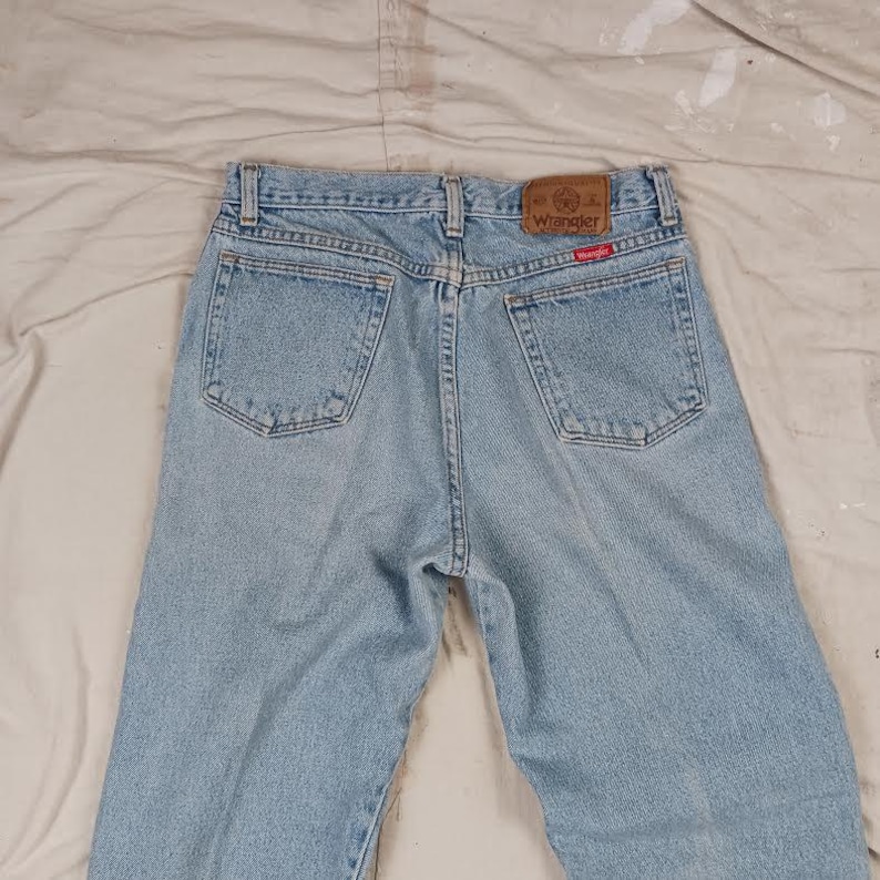 Puede incluir: Vaqueros de mezclilla azul claro con un parche de cuero marr&oacute;n que dice "Wrangler" en letras blancas. Los vaqueros tienen un estilo cl&aacute;sico de cinco bolsillos.