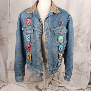 May include: A vintage denim jacket with multiple embroidered patches. The jacket has a light blue wash, a corduroy collar, and button closures. Patches include state and travel-themed designs. The jacket is displayed on a mannequin.