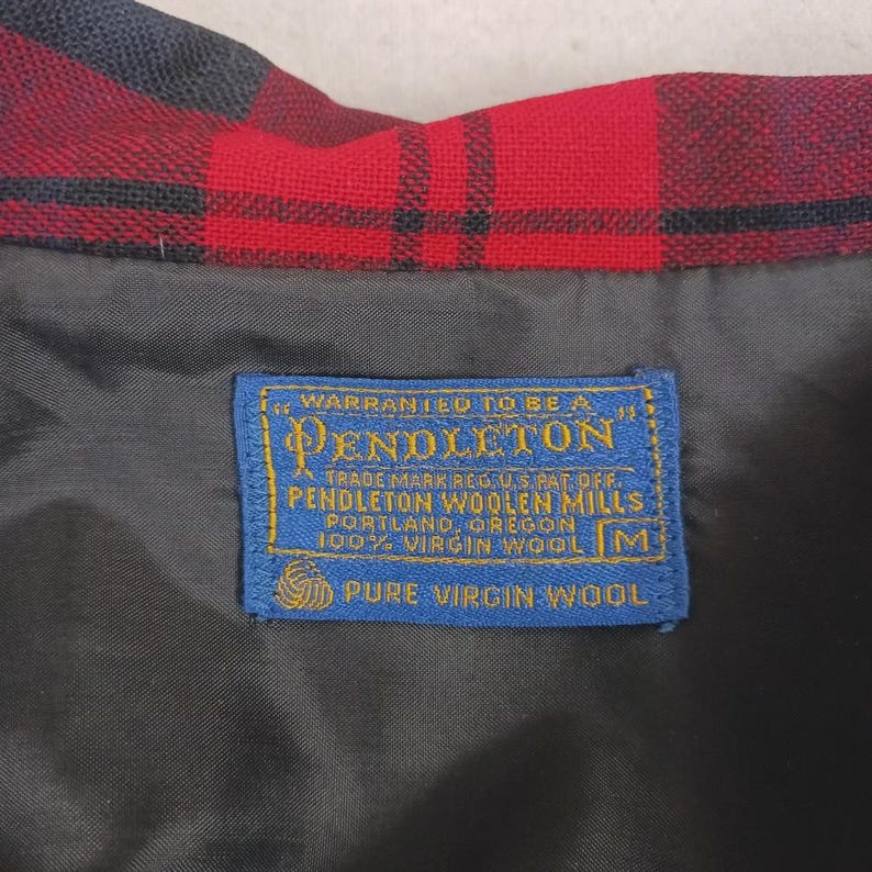 May include: Close-up of a red and black plaid fabric with a blue label. The label reads "PENDLETON WOOLEN MILLS, PORTLAND, OREGON, 100% VIRGIN WOOL". The fabric is likely wool, and the label indicates it's a quality product.