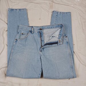 Puede incluir: Un par de pantalones vaqueros de mezclilla azul claro con cierre de bot&oacute;n y cinco bolsillos. Los pantalones est&aacute;n colocados sobre una superficie blanca.