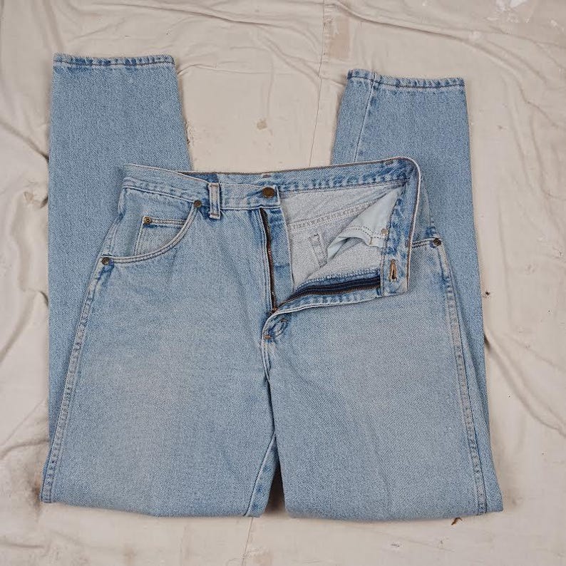 Puede incluir: Un par de pantalones vaqueros de mezclilla azul claro con cierre de bot&oacute;n y cinco bolsillos. Los pantalones est&aacute;n colocados sobre una superficie blanca.