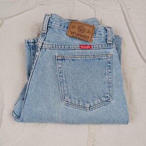 Puede incluir: Vaqueros de mezclilla azul claro con un parche de cuero marr&oacute;n que dice "Wrangler Authentic Jeans" y una etiqueta roja que dice "Wrangler".