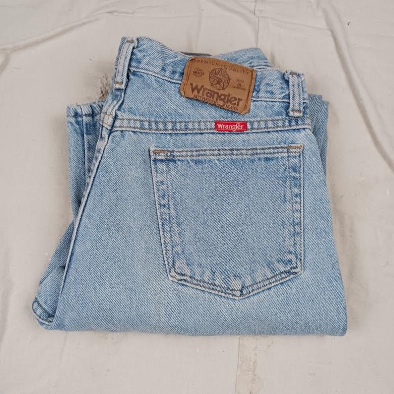 Puede incluir: Vaqueros de mezclilla azul claro con un parche de cuero marr&oacute;n que dice "Wrangler Authentic Jeans" y una etiqueta roja que dice "Wrangler".