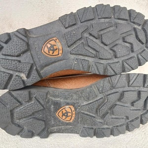 May include: Close-up of the brown leather soles of a pair of brown leather boots. The soles have a deep tread pattern and the Ariat logo is visible on both soles.