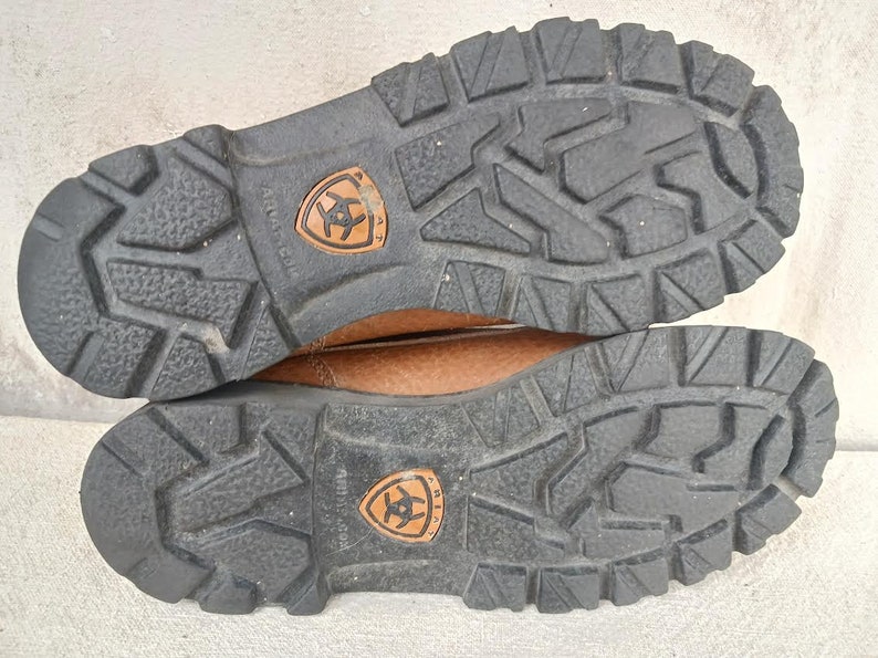 May include: Close-up of the brown leather soles of a pair of brown leather boots. The soles have a deep tread pattern and the Ariat logo is visible on both soles.