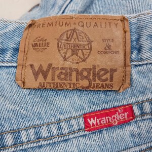 Puede incluir: Un primer plano de un jean de mezclilla azul claro con una etiqueta Wrangler de cuero marr&oacute;n que dice "Premium Quality", "Extra Value", "Wrangler Authentic Jeans", y "Style & Comfort". La etiqueta tambi&eacute;n presenta una estrella con el texto "Wrangler Authentic" en su interior. Hay una etiqueta Wrangler roja en la esquina inferior derecha del jean.