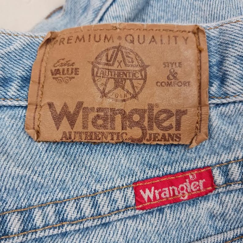 Puede incluir: Un primer plano de un jean de mezclilla azul claro con una etiqueta Wrangler de cuero marr&oacute;n que dice "Premium Quality", "Extra Value", "Wrangler Authentic Jeans", y "Style & Comfort". La etiqueta tambi&eacute;n presenta una estrella con el texto "Wrangler Authentic" en su interior. Hay una etiqueta Wrangler roja en la esquina inferior derecha del jean.