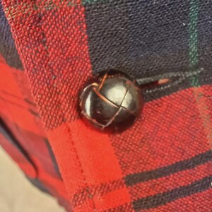 May include: Close-up of a black, round button on a red, black, and green plaid fabric. The button is textured and appears to be leather or a similar material. The plaid pattern is a classic design.