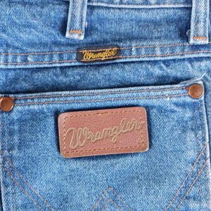 May include: Close-up of a pair of blue denim jeans with a brown leather patch that says "Wrangler" in gold lettering. The jeans have a back pocket with a stitched design.