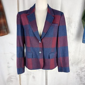 May include: A cropped, plaid blazer with a blue and burgundy checkered pattern. The blazer has a single-breasted closure with two buttons and two flap pockets.