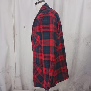 May include: Red and navy blue plaid flannel shirt with a button-down front, a chest pocket, and long sleeves. The shirt is displayed on a mannequin. The fabric has a classic lumberjack style pattern.