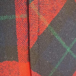 May include: Close-up of a woven fabric with a red, black, and green plaid pattern. The fabric has a textured appearance, with the red sections dominating the view. Green lines intersect the black sections.