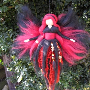 Könnte beinhalten: Eine rote und schwarze Filz-Feenpuppe mit langen, fließenden Haaren und Flügeln. Die Puppe trägt ein schwarzes Kleid mit einem roten Gürtel und eine Perlenkette mit Totenkopf-Anhängern.