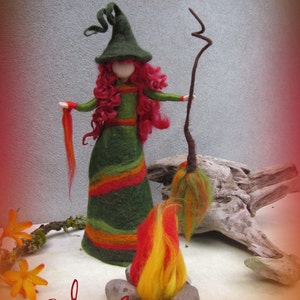 May include: A felt witch doll with long red hair, wearing a green and orange striped dress and a green pointed hat. She is holding a brown stick with a green and orange felt broom attached to the end. The doll is standing on a wooden surface with a small felt fire in front of her.