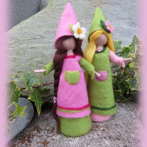 Könnte beinhalten: Zwei Filzpuppen in grünen und rosa Kleidern mit passenden Hüten. Die Puppen haben langes braunes und blondes Haar und halten Blumen.