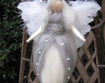 Puede incluir: Un ángel de fieltro gris y blanco con alas blancas y una estrella plateada en el pecho. El ángel está colgado de un enrejado metálico.