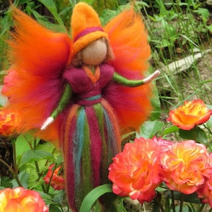 Könnte beinhalten: Eine Filz-Feenpuppe mit orange-rotem Haar, einem burgunderfarbenen Kleid mit bunten Streifen und einem leuchtend orangefarbenen Hut. Die Fee steht in einem Garten mit orangen Rosen.
