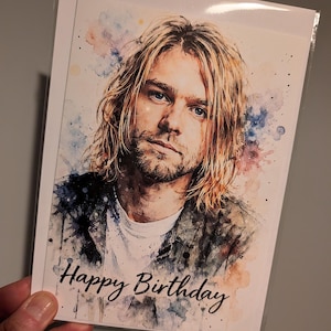 May include: A birthday card with a watercolour portrait of a man with long blonde hair and blue eyes. The card reads "Happy Birthday" in black script. The background features colourful watercolour splashes.