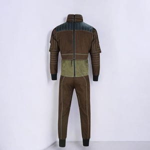 Puede incluir: Mono marr&oacute;n con cuello alto y un panel verde en la secci&oacute;n media. Los hombros y la parte superior de la espalda presentan un acento verde m&aacute;s oscuro. Las mangas tienen un dise&ntilde;o en capas y los pu&ntilde;os son negros. El pantal&oacute;n es recto.