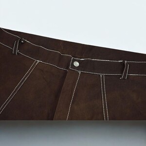 Puede incluir: Primer plano de pantalones marrones con costuras blancas. Los pantalones tienen cierre de bot&oacute;n y trabillas para el cintur&oacute;n. Las costuras blancas proporcionan un detalle de contraste a la tela marr&oacute;n. Los pantalones parecen ser de un material resistente.