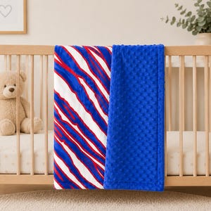 May include: A rectangular baby blanket featuring a dual-sided design. One side displays a vibrant pattern of diagonal red, white, and blue stripes. The reverse side consists of a solid royal blue fabric with a raised, textured dimpled surface.