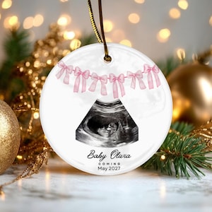 Peut inclure: Une décoration ronde en céramique blanche avec une échographie en noir et blanc d'un fœtus. Au-dessus de l'image se trouve une guirlande de nœuds roses. Sous l'image, le texte indique Baby Olivia, COMING, May 2027. L'ornement est suspendu par un cordon doré.
